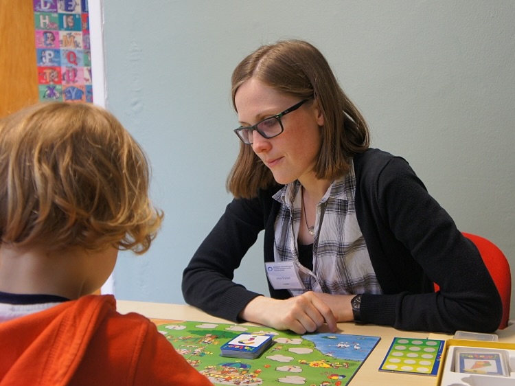 Logopädin arbeitet mit einem Kind an einem Tisch mit Lernmaterialien und Spielen zur Sprachförderung.