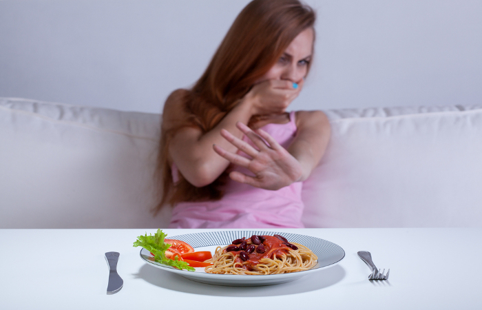 Frau sitzt vor einem Teller Pasta, wirkt angewidert und hält sich den Mund zu – Symbolbild für Ess-Brech-Sucht (Bulimie).