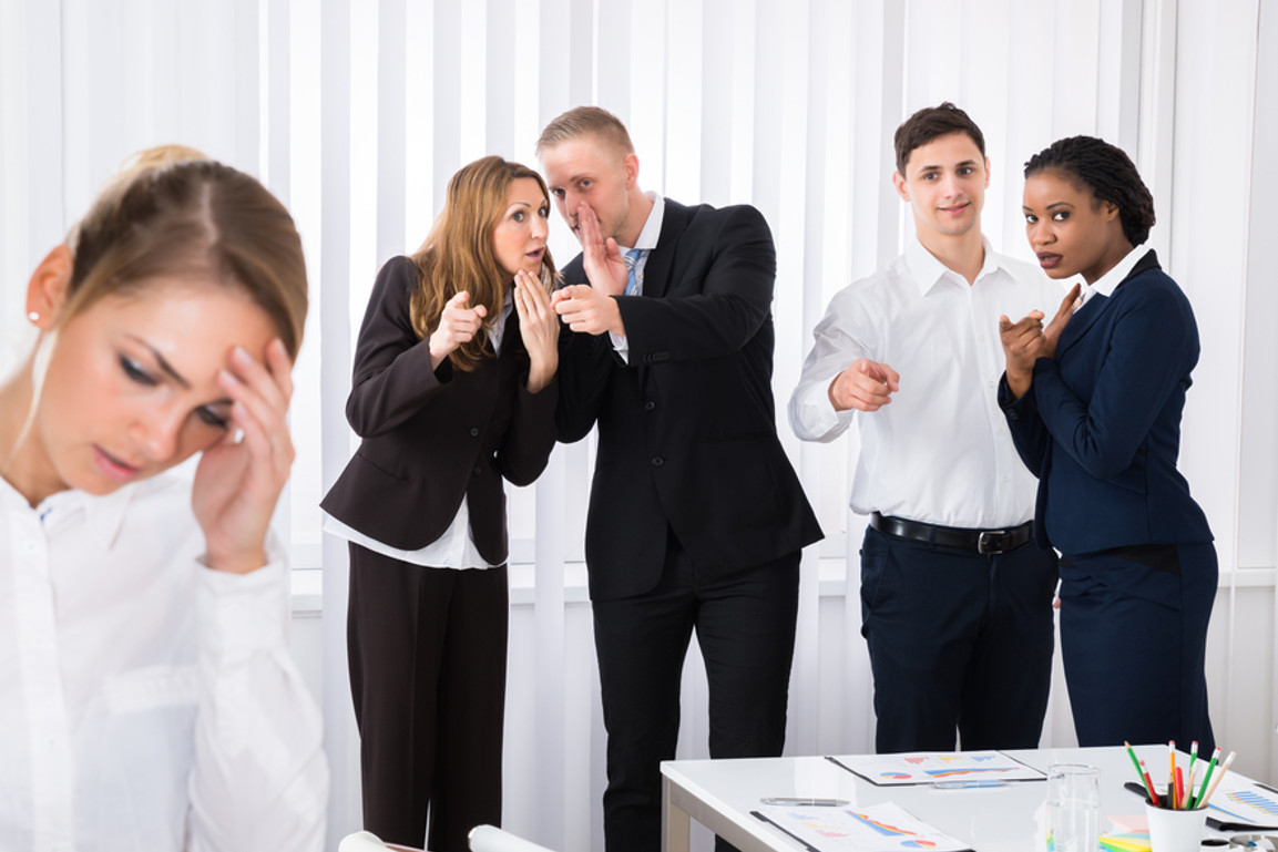Junge Frau verzweifelt am Arbeitsplatz, während Kollegen tuscheln und sie ausgrenzen – Symbolbild für Mobbing und Psychoterror.