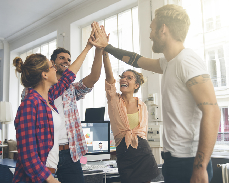 Vier junge Erwachsene geben sich im Büro ein High-Five – Symbolbild für Teamgeist