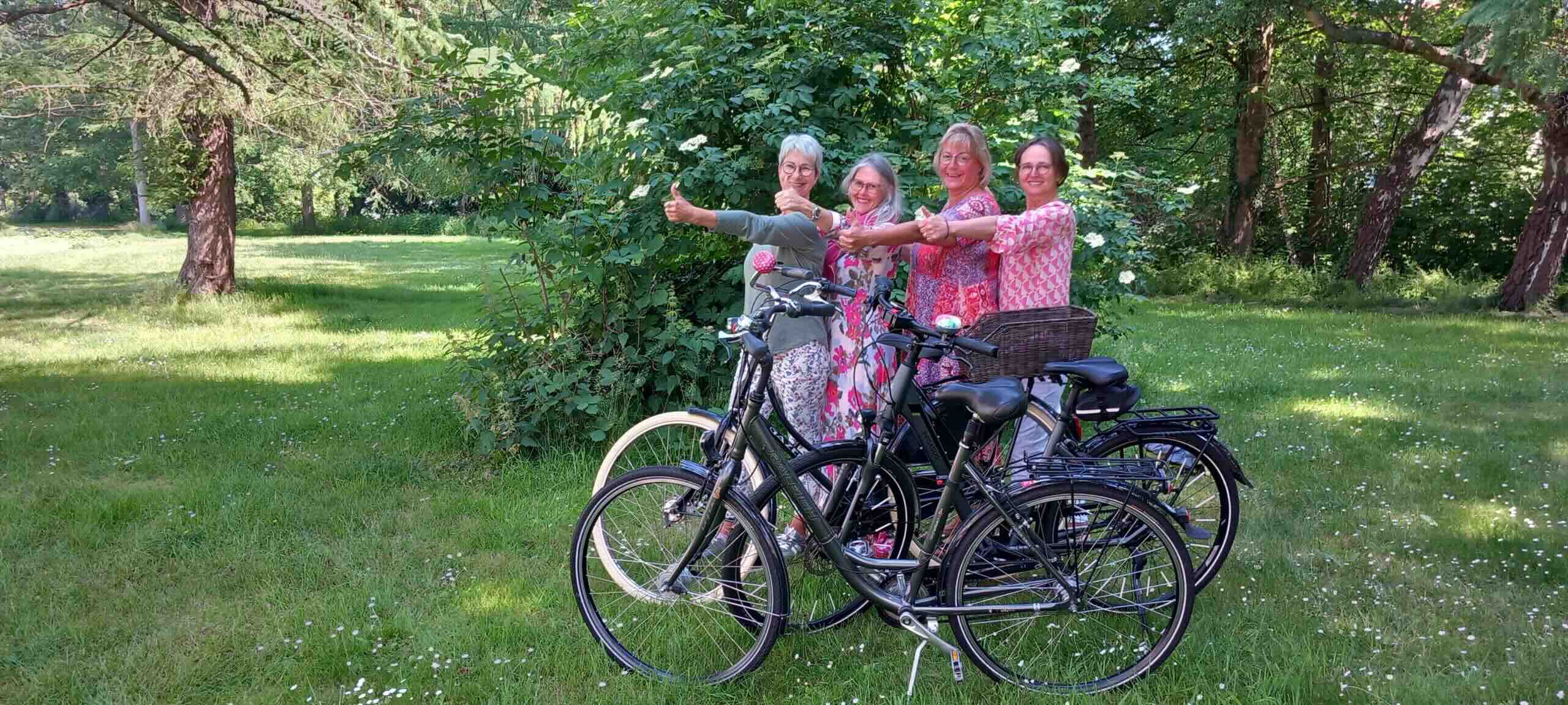 Vier Frauen stehen mit Fahrrädern nebeneinander auf einer Wiese, heben gemeinsam den Daumen hoch und lächeln in die Kamera.