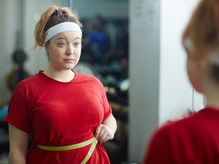 Junge Frau in rotem T-Shirt mit Maßband um die Taille schaut in den Spiegel – Symbolbild für Übergewicht, Body Mass Index (BMI) und Essverhalten.